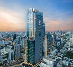 A tall, modern glass skyscraper with the Chatrium Grand logo stands prominently among other buildings in a bustling urban cityscape at sunset. The sky glows with warm pastel colors.
