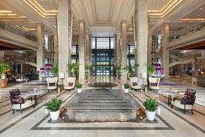 Spacious, elegant hotel lobby with high ceilings, large windows, marble floors, central water feature, potted plants, and seating areas with modern chairs and tables arranged symmetrically.