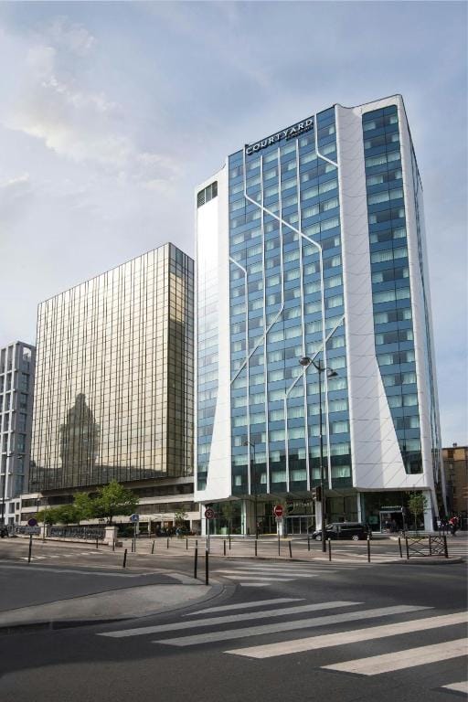 Modern glass buildings, including a tall white and blue Courtyard hotel, stand next to a street with crosswalks, traffic signs, and some greenery in an urban setting under a partly cloudy sky.
