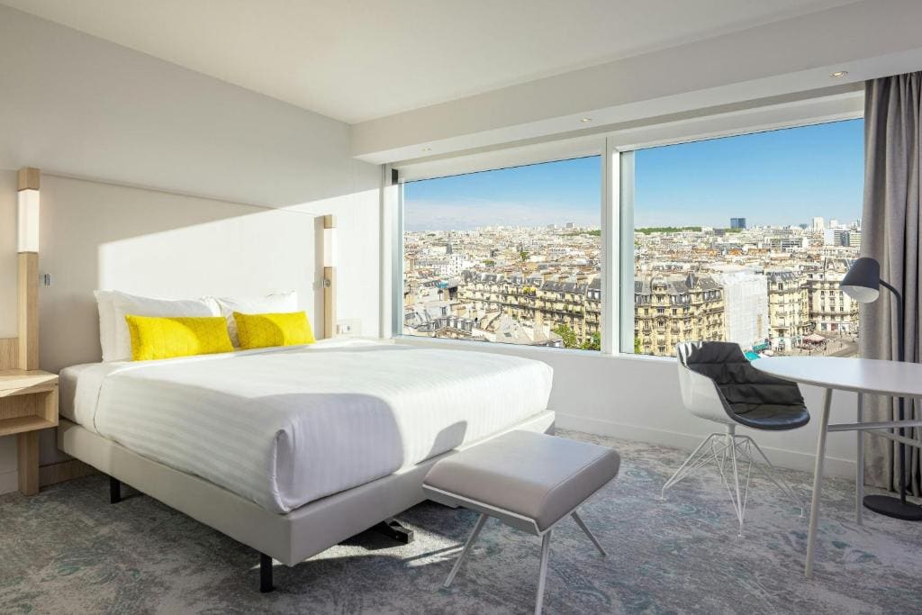 A modern hotel room with a large bed, white linens, yellow cushions, a desk, and a chair. Floor-to-ceiling windows offer a bright view of Paris city rooftops and buildings under a clear sky.