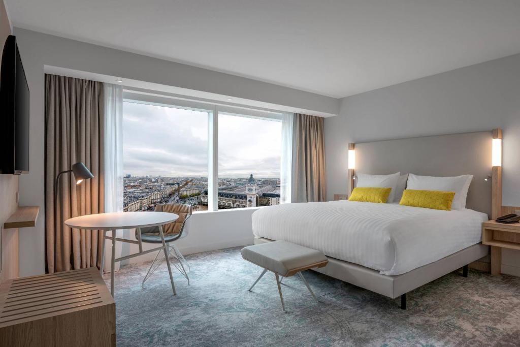 A modern hotel room with a large bed, white linens, two yellow accent pillows, a bench, a round table with two chairs, a floor lamp, and a large window offering a city view. Neutral tones and natural light fill the space.