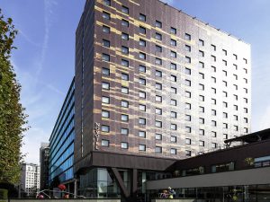 A modern multi-story hotel building with a geometric facade pattern, large windows, and the word “Novotel” displayed vertically near the entrance, set against a blue sky. Trees line the left side of the image.