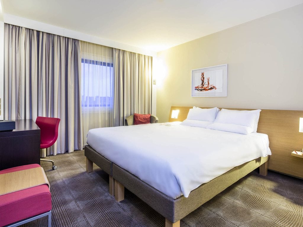Modern hotel room with a large bed, white linens, bedside tables, a desk with a red chair, armchair, window with curtains, and a framed artwork on the wall above the bed.