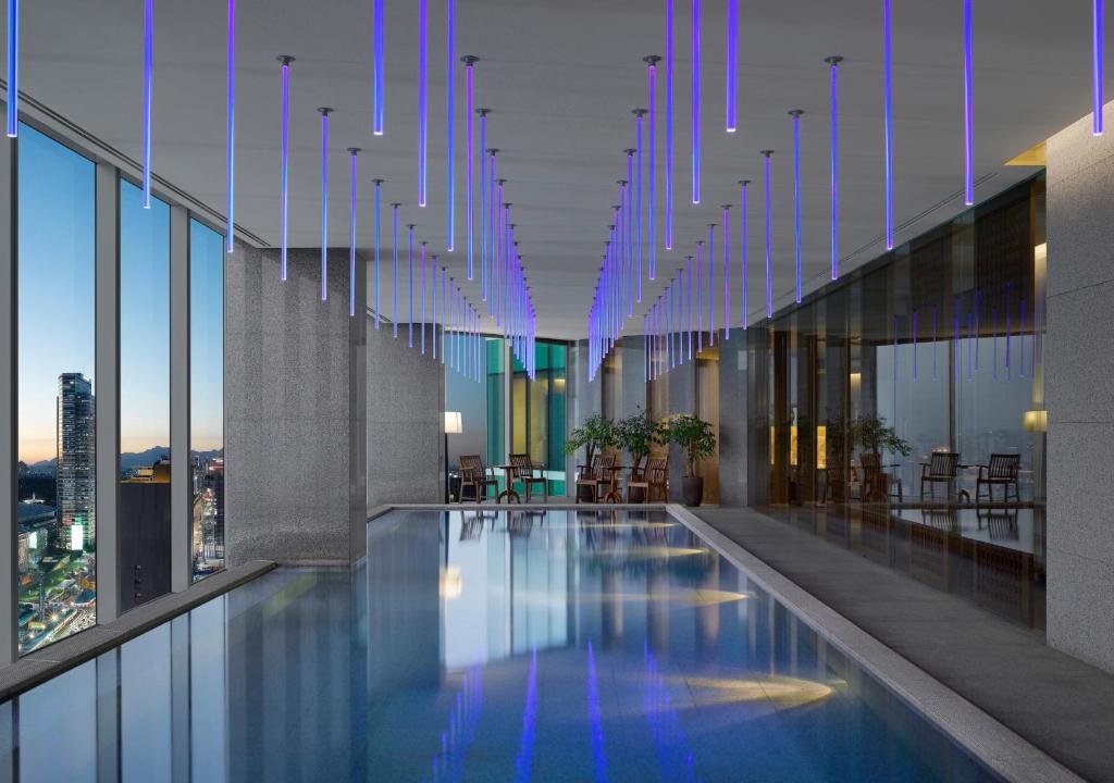 Indoor pool with blue hanging lights on the ceiling, floor-to-ceiling windows showing a cityscape at dusk, and a seating area with tables and plants along one side.