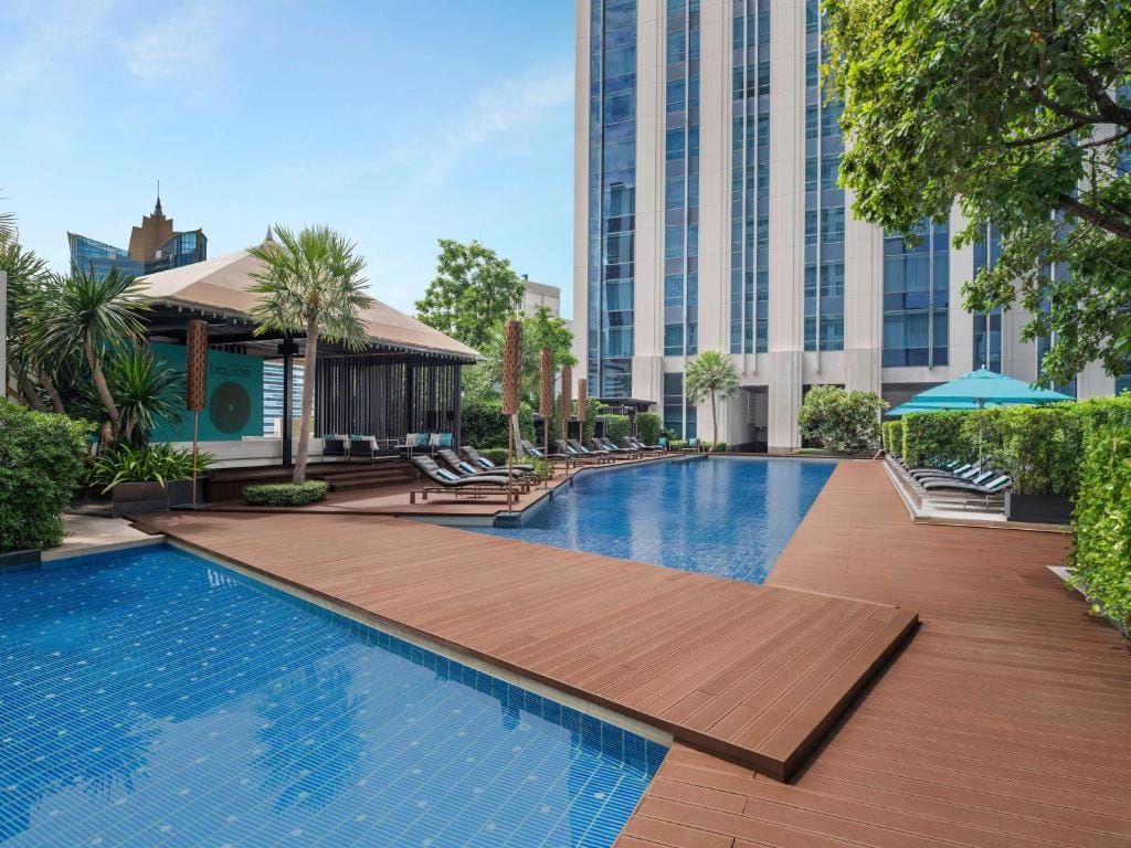 A modern outdoor pool area with lounge chairs, umbrellas, palm trees, and a wooden deck, surrounded by tall buildings and greenery on a sunny day.