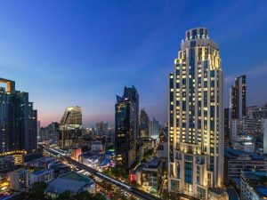 Cityscape at dusk with tall, illuminated buildings, including a prominent hotel labeled SOFITEL, roads with light trails from traffic, and a vibrant blue sky transitioning to night.