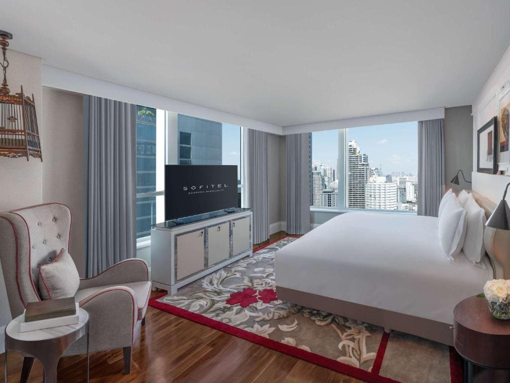 Modern hotel room with large windows offering city views, a king-size bed, gray armchair, flat-screen TV on a cabinet, floral rug, and neutral-toned décor. Natural light fills the space.