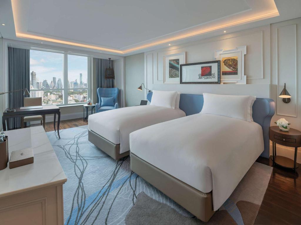 A modern hotel room with two neatly made twin beds, a large window with a city view, a desk, a blue armchair, wall art above the beds, and soft ambient lighting.