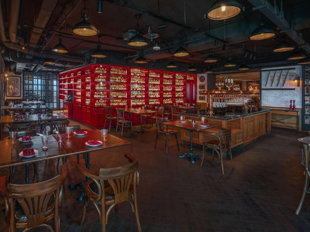 Spacious bar with wooden tables and chairs, a large bar counter, and a striking red wall filled with bottles. Warm lighting and industrial-style ceiling create a cozy, inviting atmosphere.