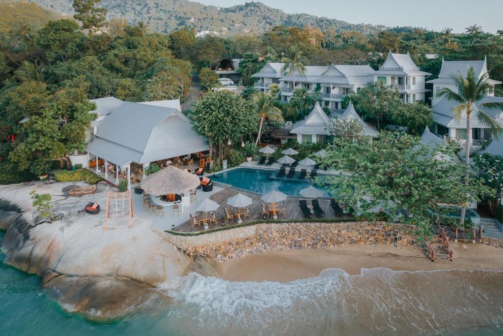 Aerial view of a beachfront resort with white buildings, a swimming pool, palm trees, lounge chairs, and a sandy beach beside turquoise ocean water, surrounded by lush greenery and hills.