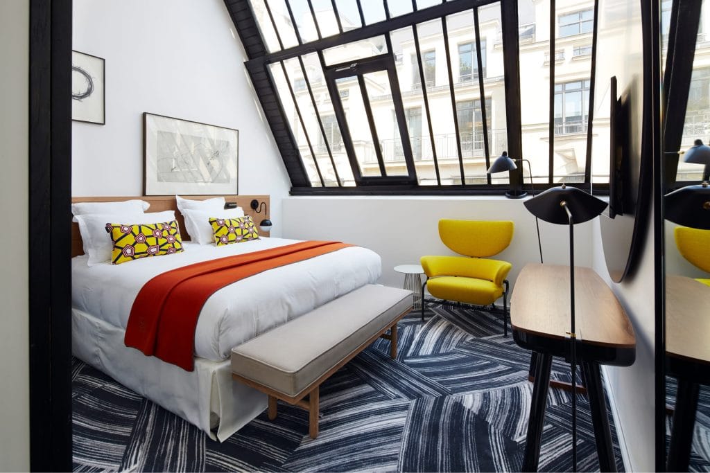 A modern hotel room with a large bed, colorful pillows, an orange throw, a yellow chair, geometric-patterned carpet, and a slanted glass wall with black frames letting in natural light.