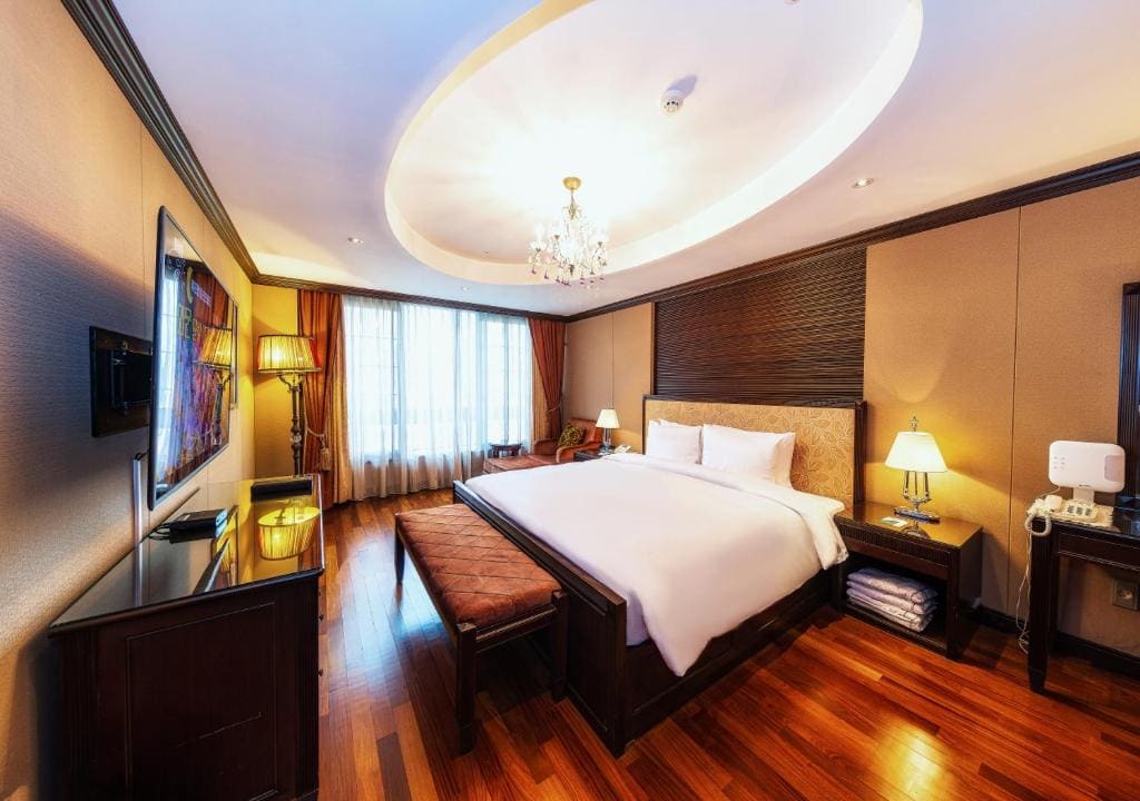 A spacious hotel room with a large bed, wooden floors, elegant furniture, a chandelier, lamps, flat-screen TV, large windows with curtains, and warm ambient lighting.