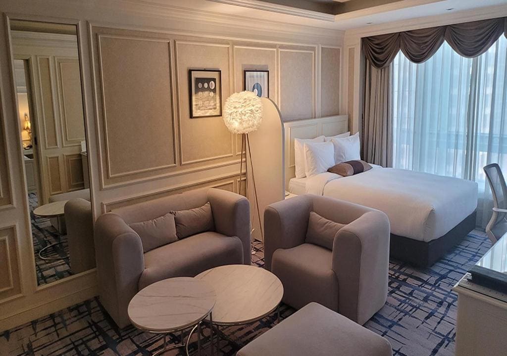 A modern hotel room with a double bed, two light gray armchairs, a small round table, a standing lamp, large window with curtains, and framed art on beige paneled walls.