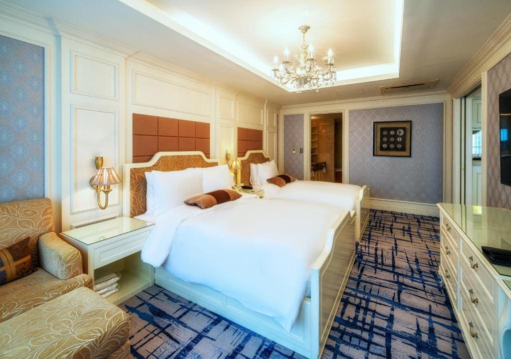 A luxurious hotel room features two large beds with white bedding, plush headboards, a chandelier, elegant furniture, patterned carpet, and soft lighting creating a cozy, upscale atmosphere.