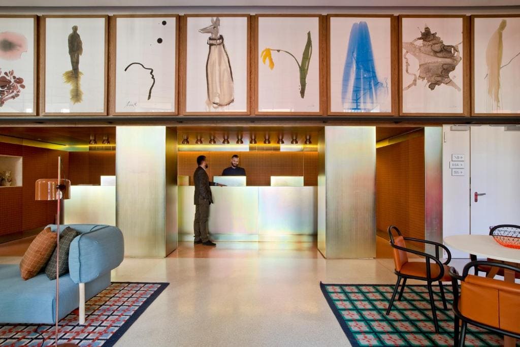 A modern hotel lobby with two people at a metallic reception desk, colorful abstract artwork above, stylish furniture, and warm lighting.