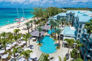 Aerial view of a beachfront resort with a turquoise pool, palm trees, and lounge chairs, adjacent to white sandy beach and clear blue ocean with sailboats and water activities. Modern buildings with blue roofs surround the pool.