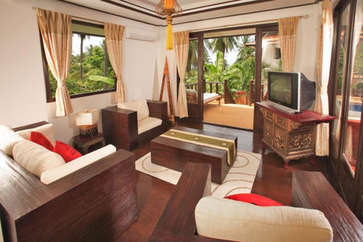 A bright, tropical-themed living room with wooden furniture, cream cushions, red accent pillows, large windows, a TV on a carved stand, and doors opening to a balcony with lush green views outside.
