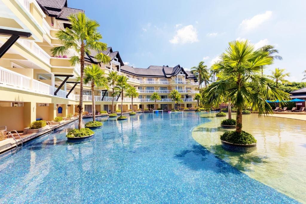 A large resort pool surrounded by palm trees and yellow, multi-story hotel buildings with balconies; clear blue sky and bright sunlight create a relaxing tropical atmosphere.