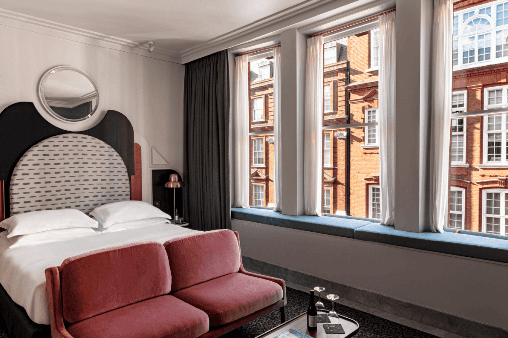 A stylish hotel room with a modern double bed, a pink sofa at the foot, large windows with black curtains, and a view of a red-brick building outside.