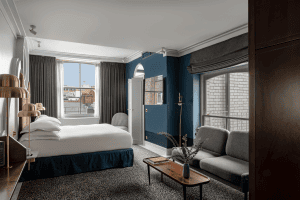 Bright, modern hotel room with a large bed, grey armchairs, a wooden coffee table, and large windows with city views. Blue accent wall, gold lamps, and neutral décor create a stylish, inviting atmosphere.