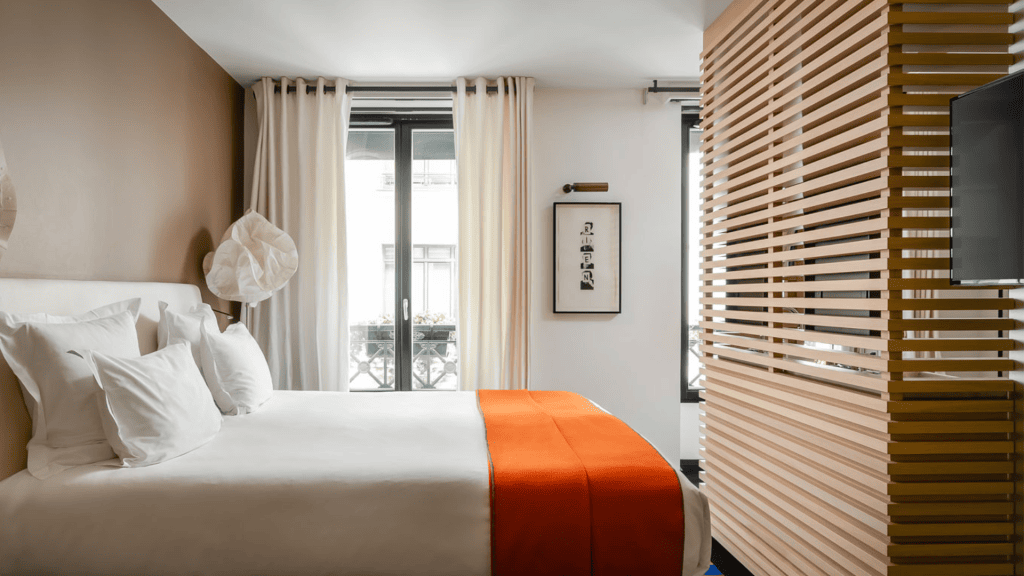 Bright, modern hotel room with a large bed dressed in white linens and an orange throw, white curtains, artwork on the wall, and a wooden slatted partition beside a wall-mounted TV.