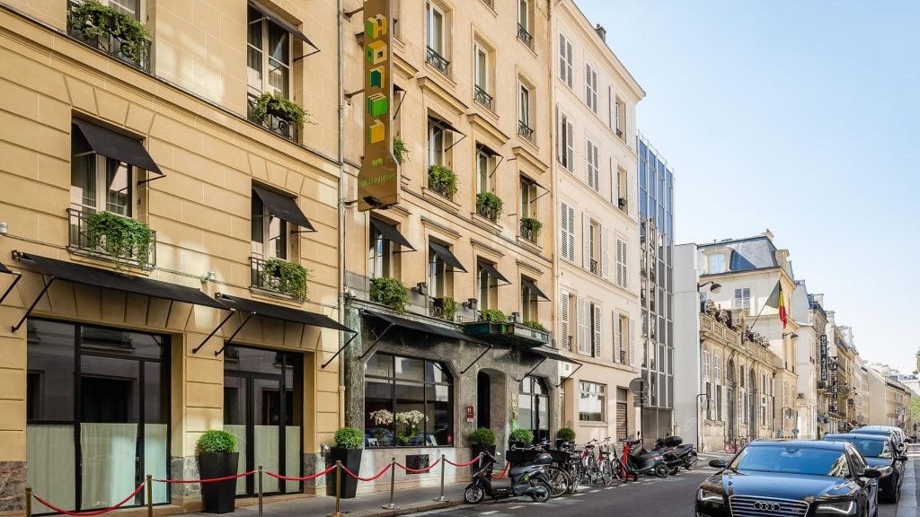 A street view of a beige-colored hotel building with black awnings and green plants on balconies. Motorcycles and cars are parked along the sidewalk, and modern glass buildings stand in the background.