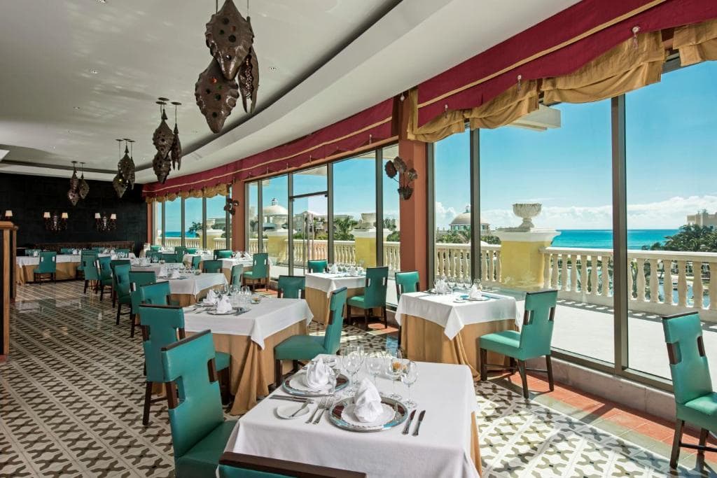 Elegant restaurant with teal chairs and white tablecloths, large windows offering a bright view of a terrace and ocean. Patterned tile floor and decorative hanging lights enhance the upscale, airy atmosphere.