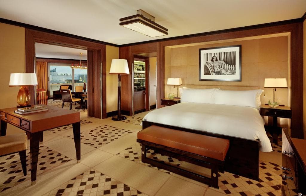 Luxurious hotel room with a large bed, two bedside tables with lamps, a bench at the foot of the bed, a desk, and an open doorway leading to a dining area with a view of the city skyline through the window.