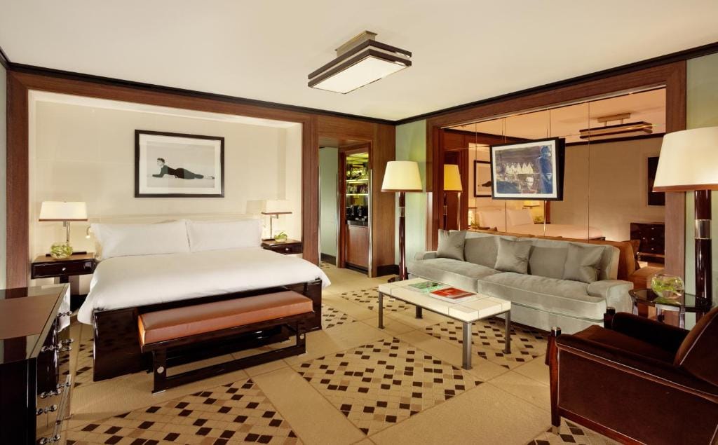Spacious hotel room with a large bed, a gray sofa, a coffee table, lamps, and decorative wall art. The room features modern decor, neutral tones, and patterned carpet flooring.