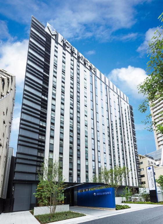 A tall, modern hotel building with HOTEL MYSTAYS signage at the top, set against a blue sky with scattered clouds, surrounded by trees and nearby buildings.