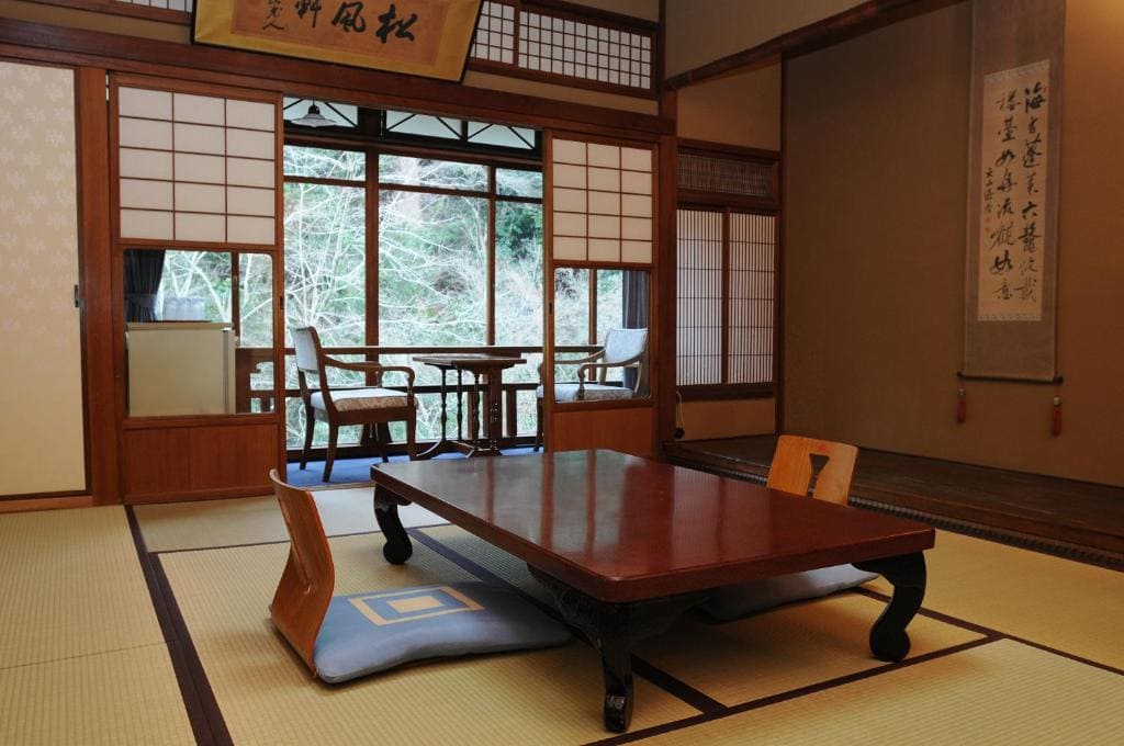 A traditional Japanese tatami room with a low wooden table, floor cushions, shoji sliding doors, and a small table with two chairs near large windows overlooking trees. Japanese calligraphy scrolls hang on the walls.