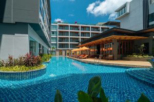 A modern hotel with a large outdoor swimming pool surrounded by lounge chairs and orange umbrellas, lush plants, and multi-story buildings under a bright blue sky with a few clouds.