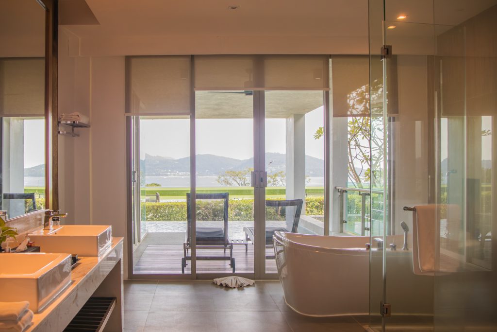 Modern bathroom with a freestanding bathtub and double sinks, featuring large glass doors that open to a patio with lounge chairs and a scenic view of mountains and water.
