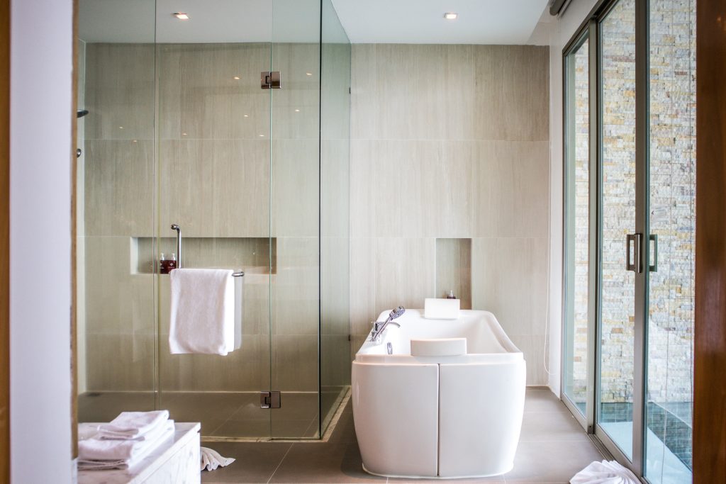 Modern bathroom with a glass-enclosed shower, a rectangular white bathtub, folded towels on a small table, and large glass doors letting in natural light. The space has light-colored tiles and a minimalist design.