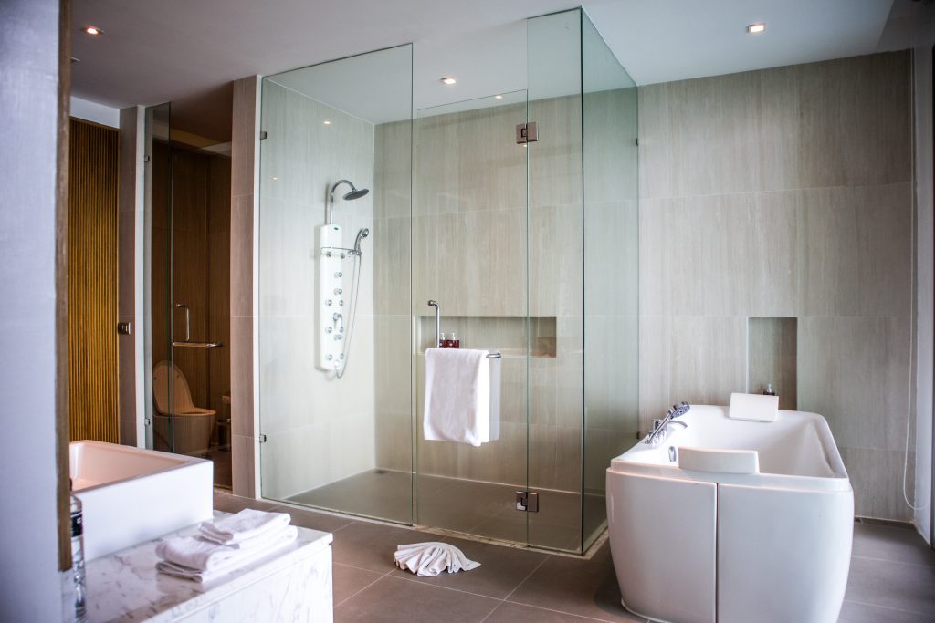 Modern bathroom with a glass-enclosed shower, a white freestanding bathtub, a sink with folded towels, and a separate toilet area. Light-colored walls and floors create a clean, contemporary look.