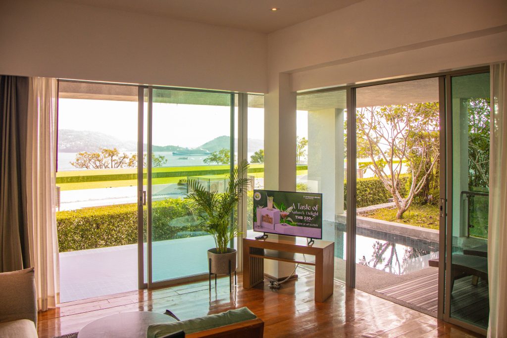 A modern living room with large glass doors opens to a terrace with a private pool, lush greenery, and a scenic view of mountains and water in the background. A TV and potted plant are placed near the sliding doors.