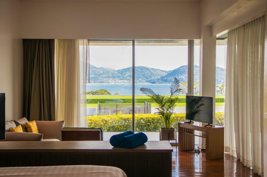 A bright hotel room with a bed, sofa, TV, and large windows showing a view of green plants, a blue sea, and distant hills under a clear sky. Two rolled blue towels sit on a wooden surface in the foreground.