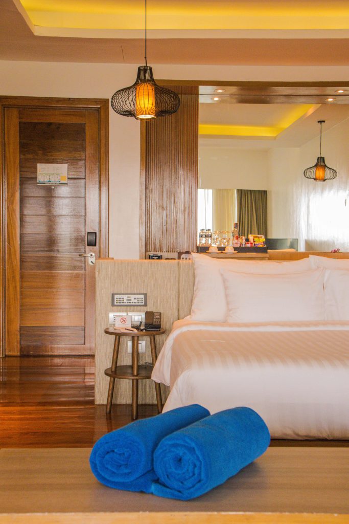 A modern hotel room with a large white bed, two blue rolled towels on a bench at the foot of the bed, wooden flooring, a bedside table with a phone, and warm ambient lighting.