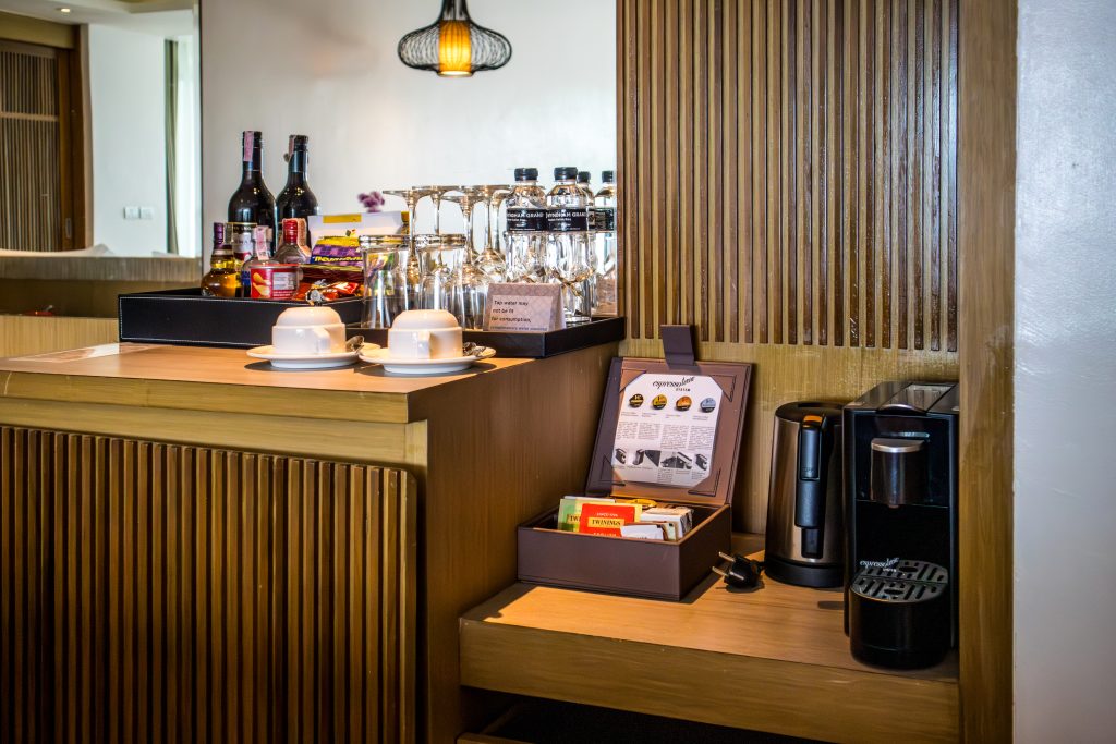 A hotel room minibar setup with bottles, glasses, snacks, two cups on saucers, a coffee machine, and a box containing tea bags and coffee supplies on a wooden counter.