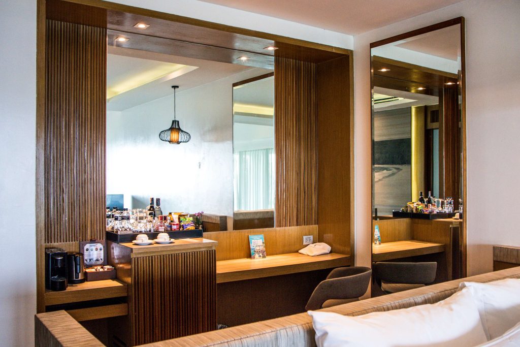 A modern hotel room features a wooden vanity with mirrors, a coffee maker, cups, bottled water, snacks, and drinks. Two chairs are placed at the desk, and soft lighting adds a cozy atmosphere.