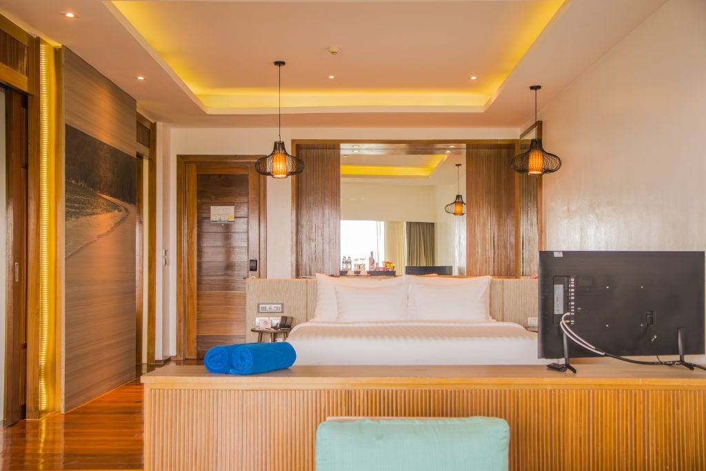 A modern hotel room with a large bed, two blue towels, wooden walls, a flat-screen TV, pendant lights, and a mirror behind the bed reflecting the room’s entrance and window.
