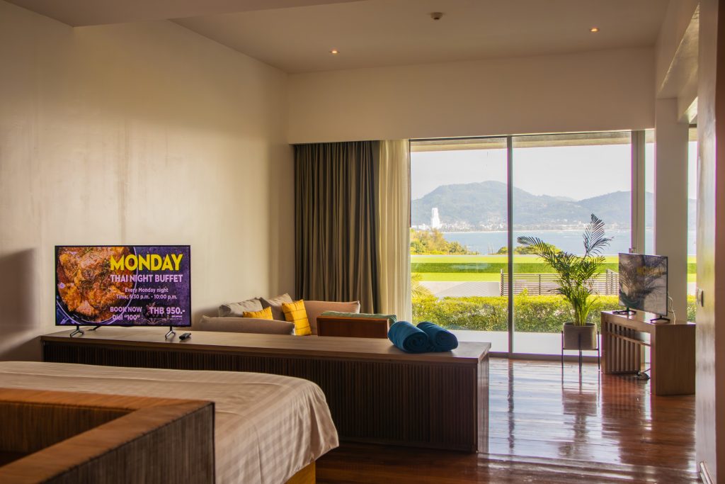 A spacious hotel room with a bed, a sofa, two TVs, and large windows revealing a scenic view of mountains and water outside. Towels are rolled on the sofa, and sunlight brightens the wooden floor.