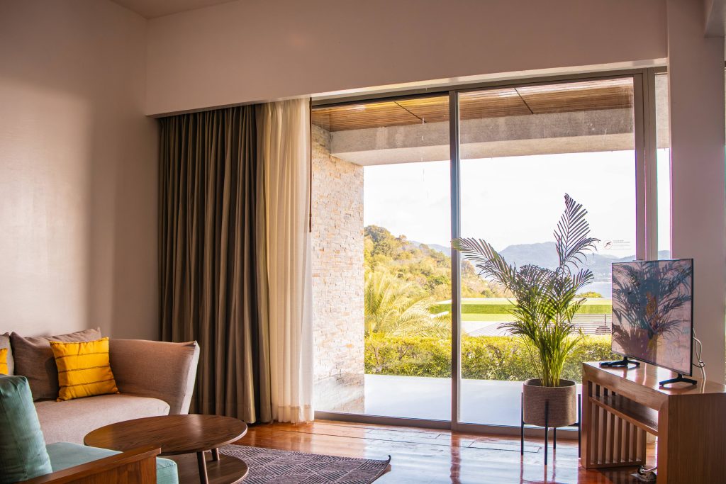 A modern living room with a sofa, coffee tables, a TV on a stand, and a large glass door opening to a balcony with plants and a scenic outdoor view of hills and greenery.
