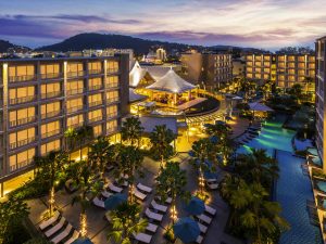 A modern resort with illuminated buildings, a large outdoor swimming pool, palm trees, lounge chairs, and a vibrant central pavilion at sunset, with hills visible in the background.