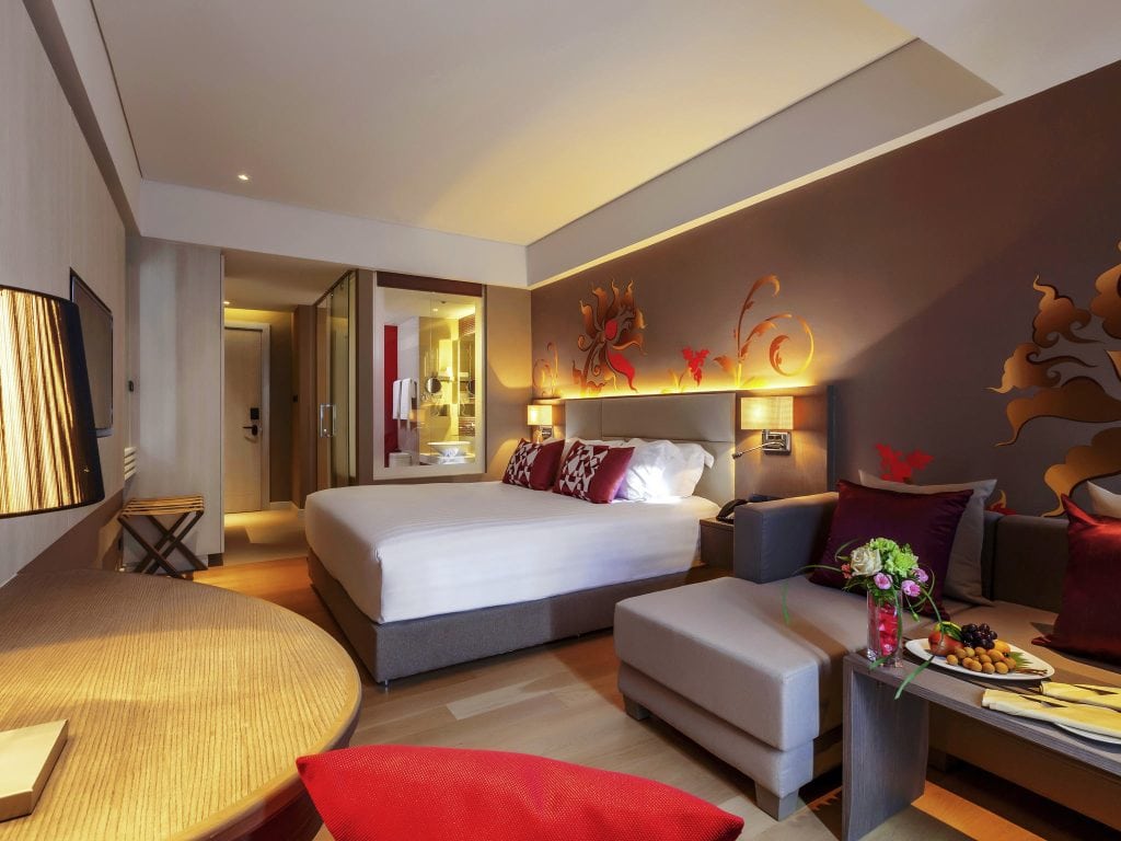 Modern hotel room with a large bed, stylish wall art, a sofa with red cushions, a table with flowers and fruit, soft lighting, and an open bathroom area in the background.