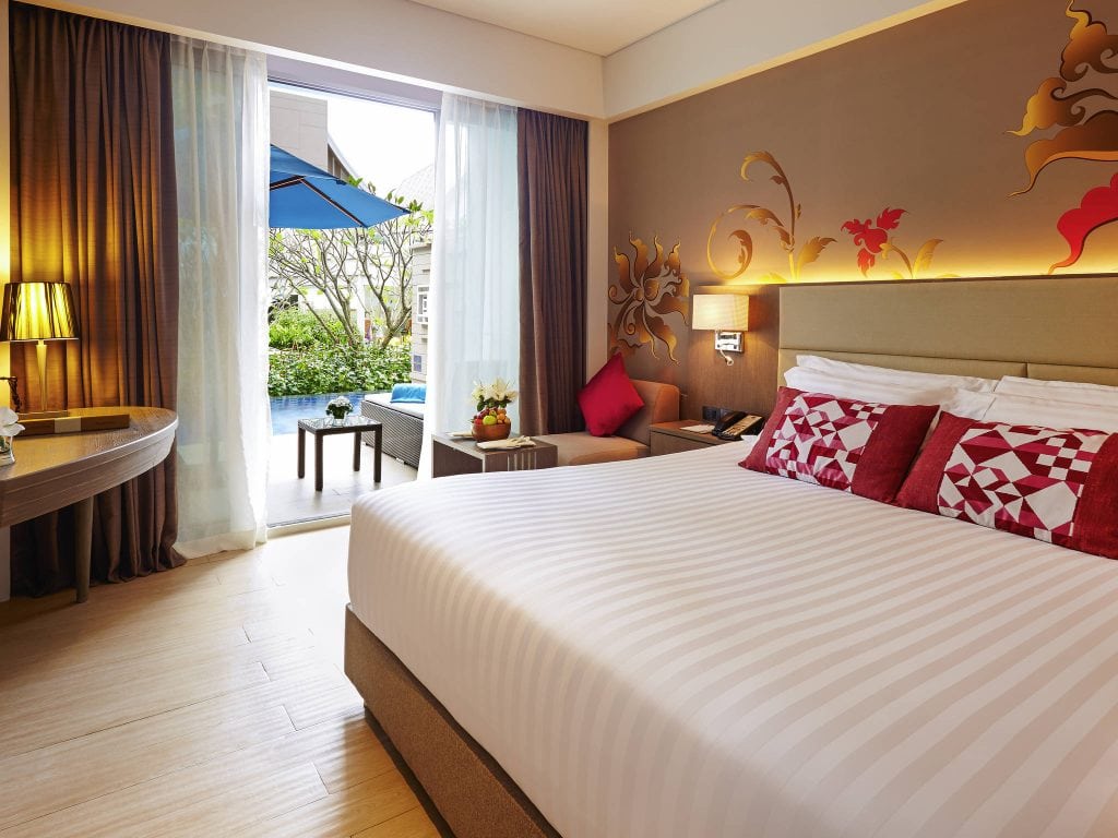 A modern hotel room with a large bed, red and white pillows, decorative wall art, a small table with a chair, and sliding glass doors leading to a patio and pool area outside.