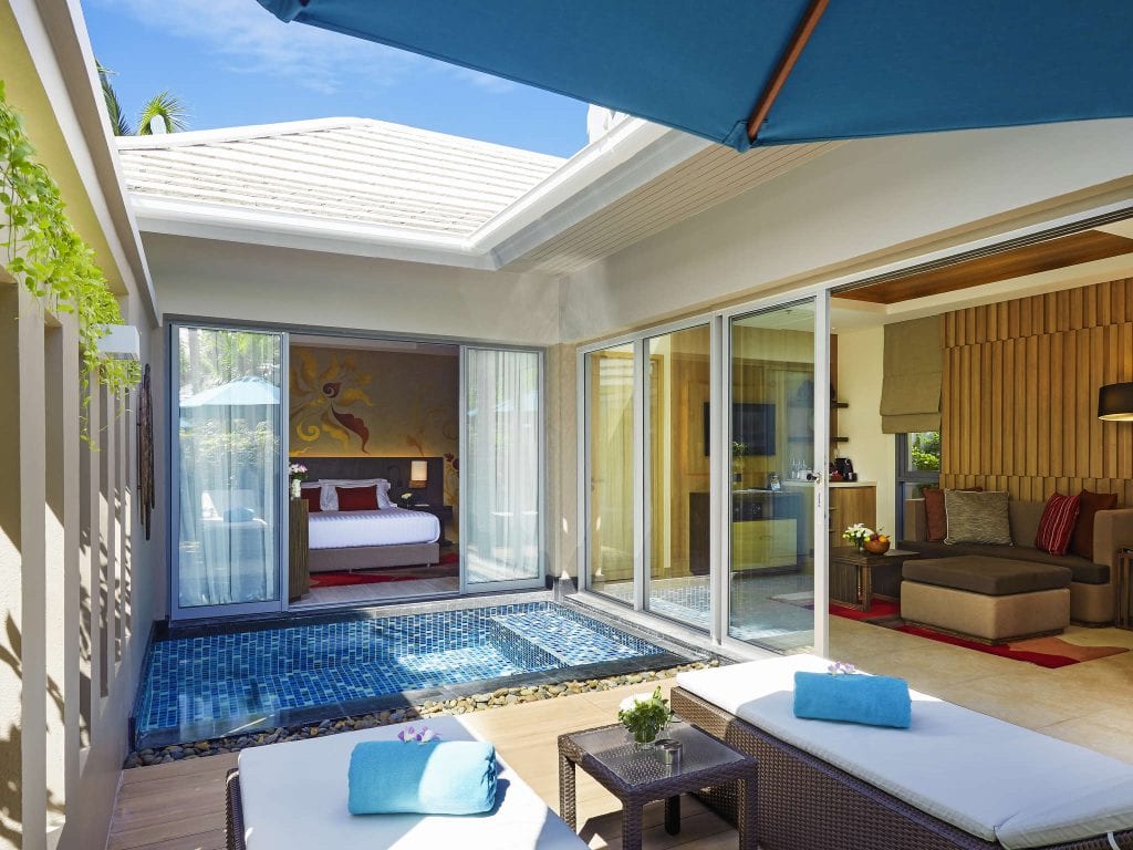 A luxurious poolside suite features lounge chairs, a small pool, and sliding glass doors leading to a bedroom with a large bed. The space is bright and modern with cozy furnishings and tropical decor.
