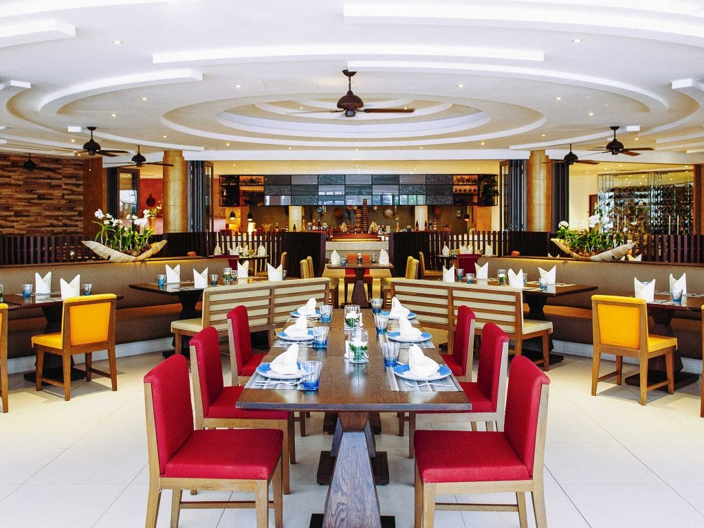 A modern, spacious restaurant interior with circular ceiling designs, red and yellow cushioned chairs, neatly set tables with napkins and glassware, and a bar area in the background.