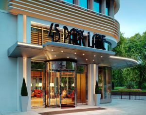The glass entrance of 45 Park Lane hotel features modern design, large black signage above revolving doors, and two potted topiary trees, with greenery and trees visible in the background.