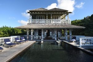 Two-story villa with a wraparound balcony, wooden roof, and white railings, featuring a rectangular pool surrounded by blue lounge chairs, set in a lush, green landscape under a blue sky with clouds.